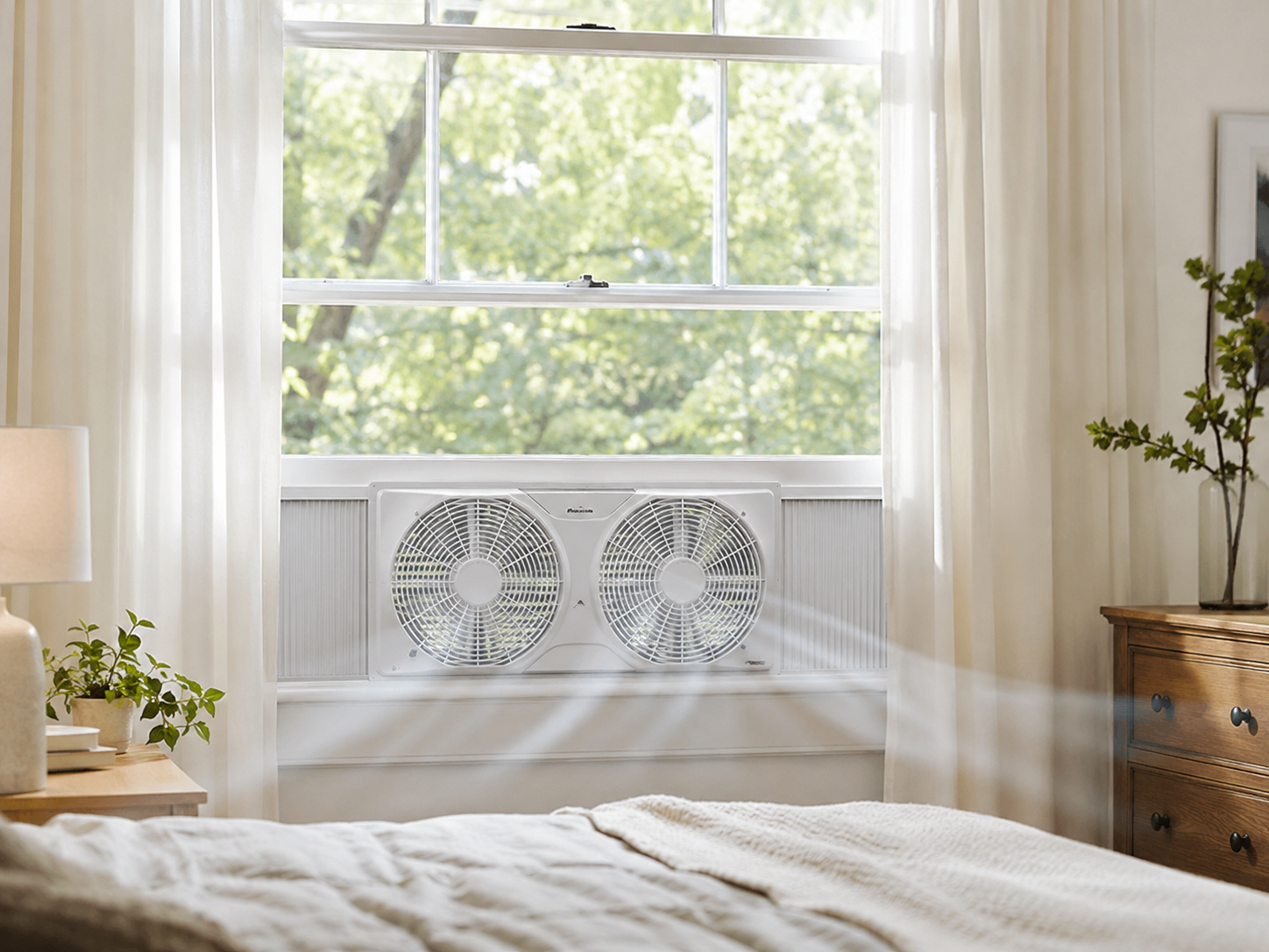 Generic twin window fan installed in a bright bedroom window with soft curtains daylight and airflow moving into the room